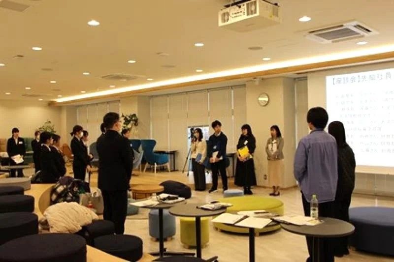 明るくモダンな空間で、学生らしき人々と社会人が集まり、プロジェクターを使った座談会やセミナーが行われている様子です。参加者は熱心に話を聞いています。